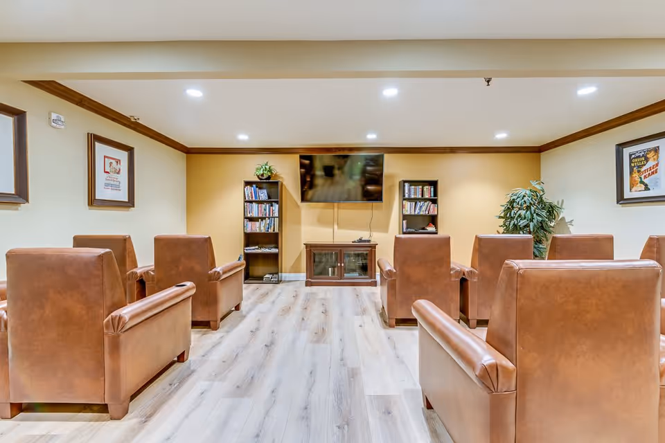 A cozy senior living common area with brown leather armchairs arranged in rows facing a wall-mounted flat-screen TV. The room has light wood flooring, two bookshelves filled with books on either side of the TV, framed artwork on the walls, and a potted plant in the corner.