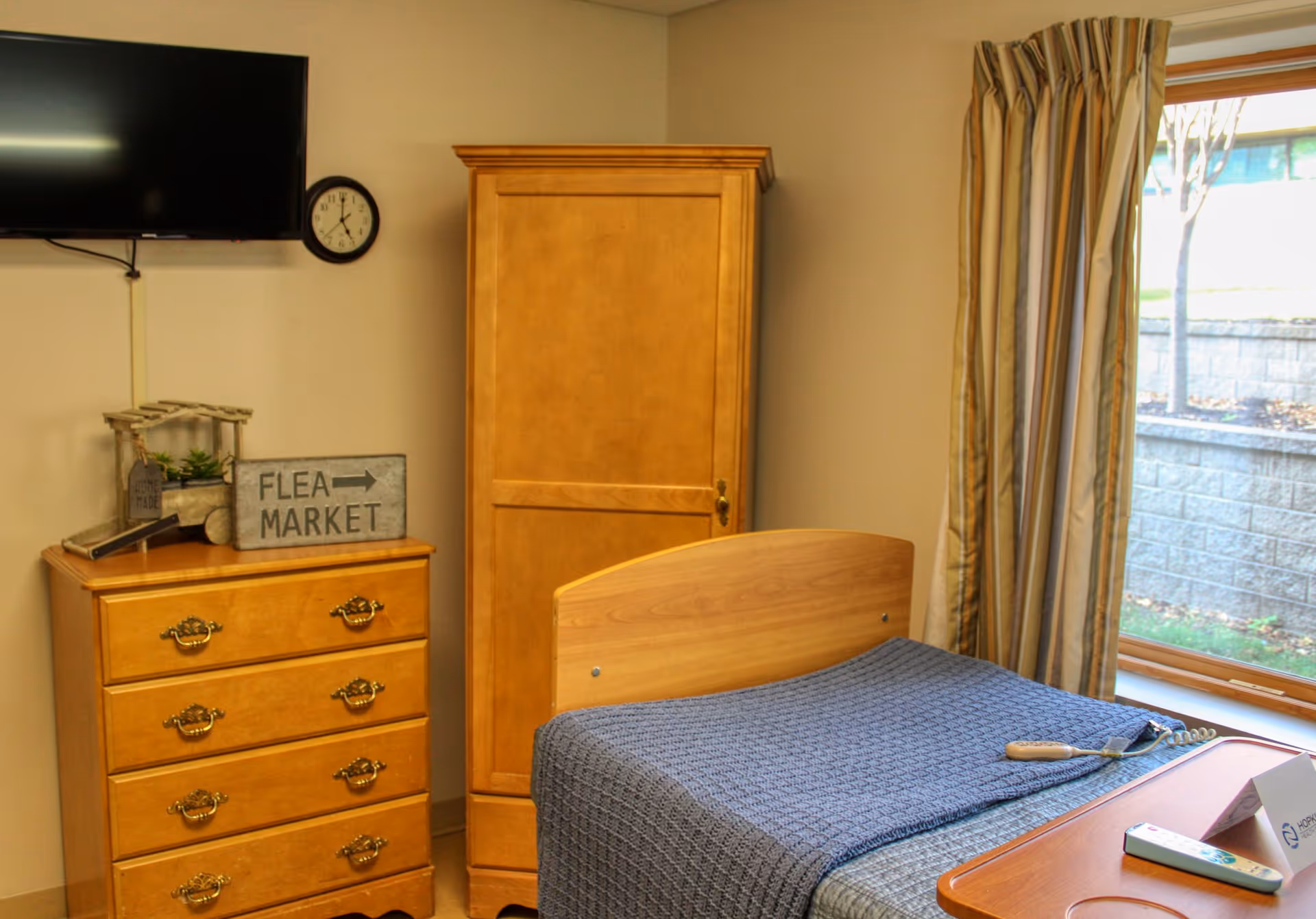 A small bedroom with a single bed covered with a blue blanket, a wooden dresser with decorative items including a sign that says 'FLEA MARKET', a wooden wardrobe, a wall-mounted TV, a clock, and a window with striped curtains showing an outdoor view.