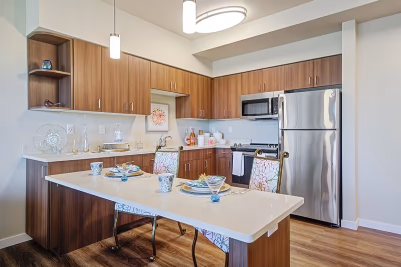 Modern kitchen with wooden cabinets, stainless steel refrigerator and microwave, a white countertop island set with plates, cups, and glasses, and two floral-patterned chairs. The kitchen has pendant and ceiling lights and wood flooring.