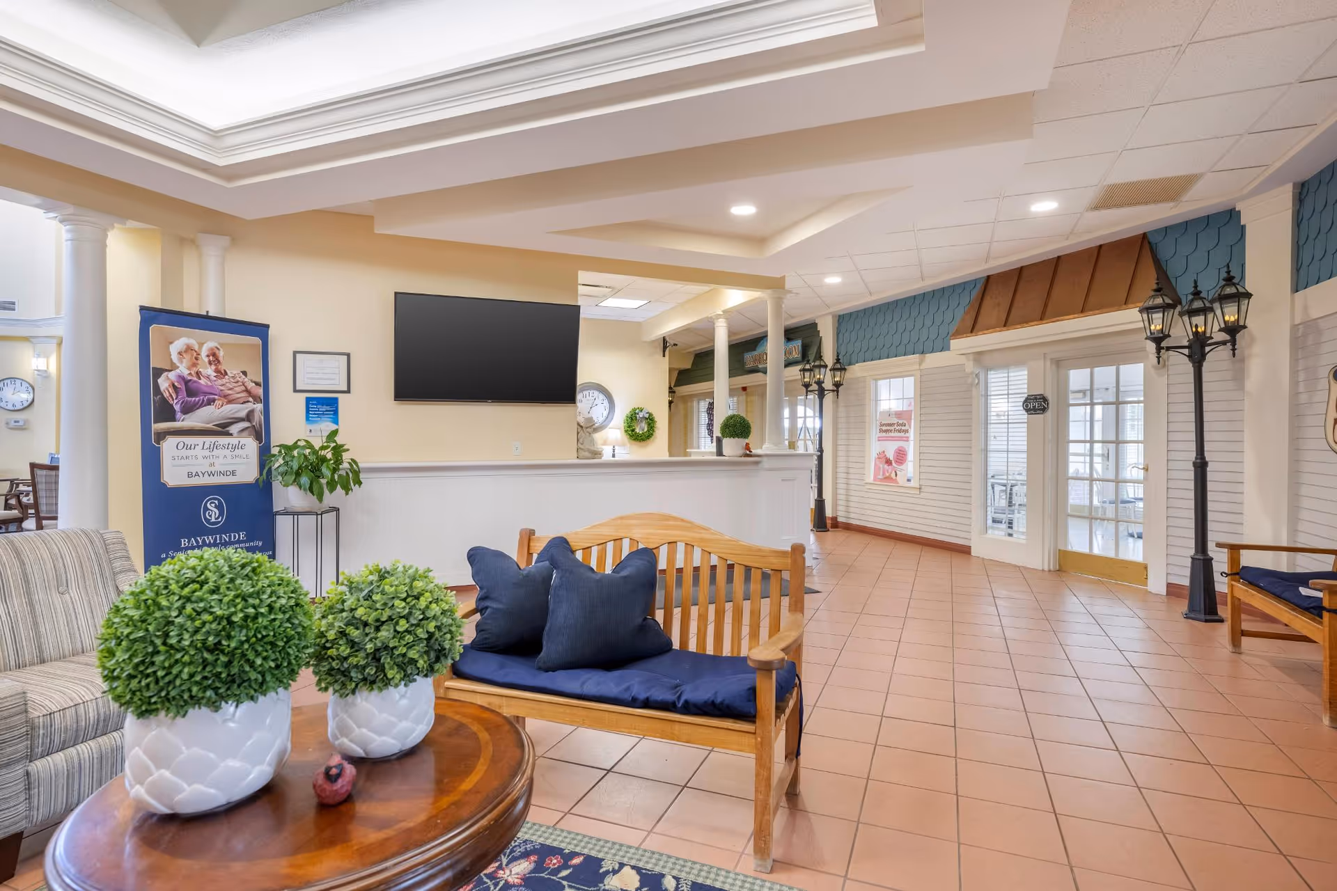 A spacious and well-lit common area in a senior living facility with tiled floors, a wooden bench with navy blue cushions, a round wooden table with two potted plants, and a striped armchair. The walls are decorated with white paneling and blue accents, and there are vintage-style street lamps indoors. A large flat-screen TV is mounted on the wall above a half-wall partition. A banner with an elderly couple and the text 'Our Lifestyle Starts With A Smile Baywinde' is visible near the seating area.