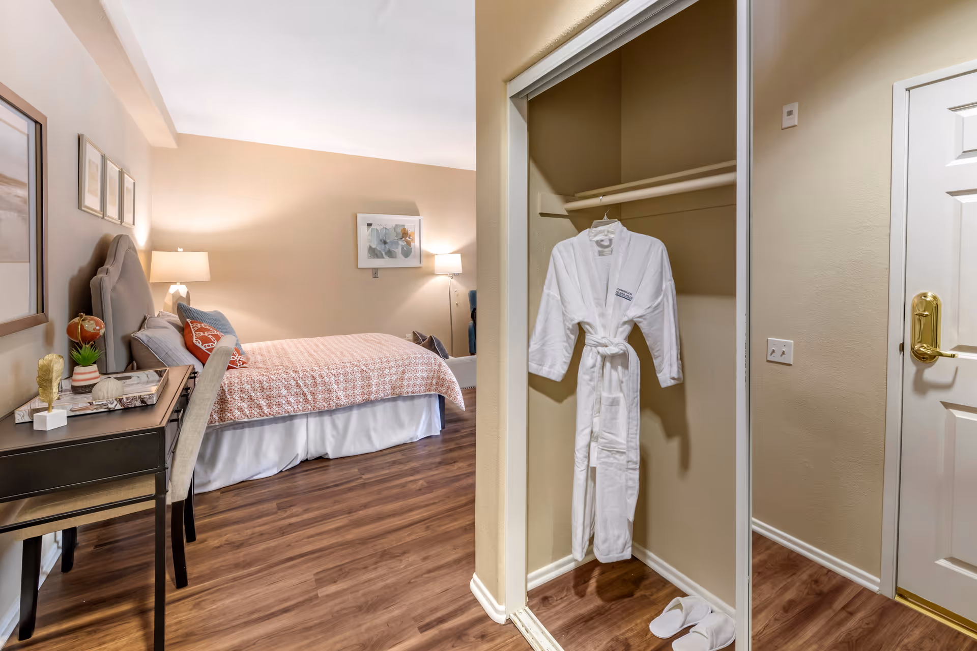 A cozy bedroom with a bed covered in a patterned bedspread, a bedside table with a lamp, and a desk with a chair. There is an open closet with a white bathrobe hanging inside and a pair of white slippers on the floor. The room has wooden flooring and beige walls with framed artwork.