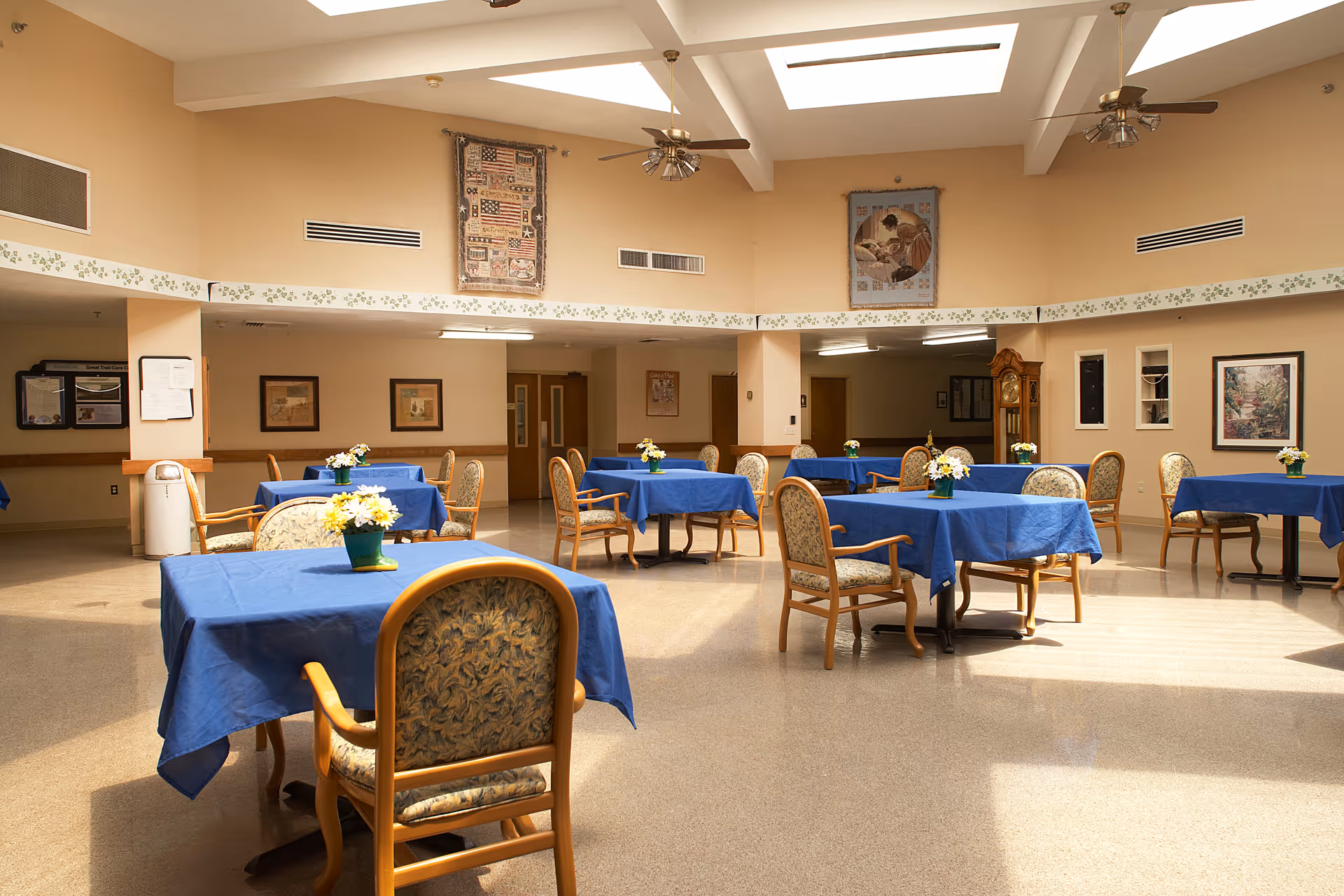 Spacious dining room with tables draped in blue tablecloths, wooden chairs, and floral centerpieces under skylights.