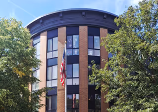 Curved brick building exterior with tall windows, an American flag, and trees in front.