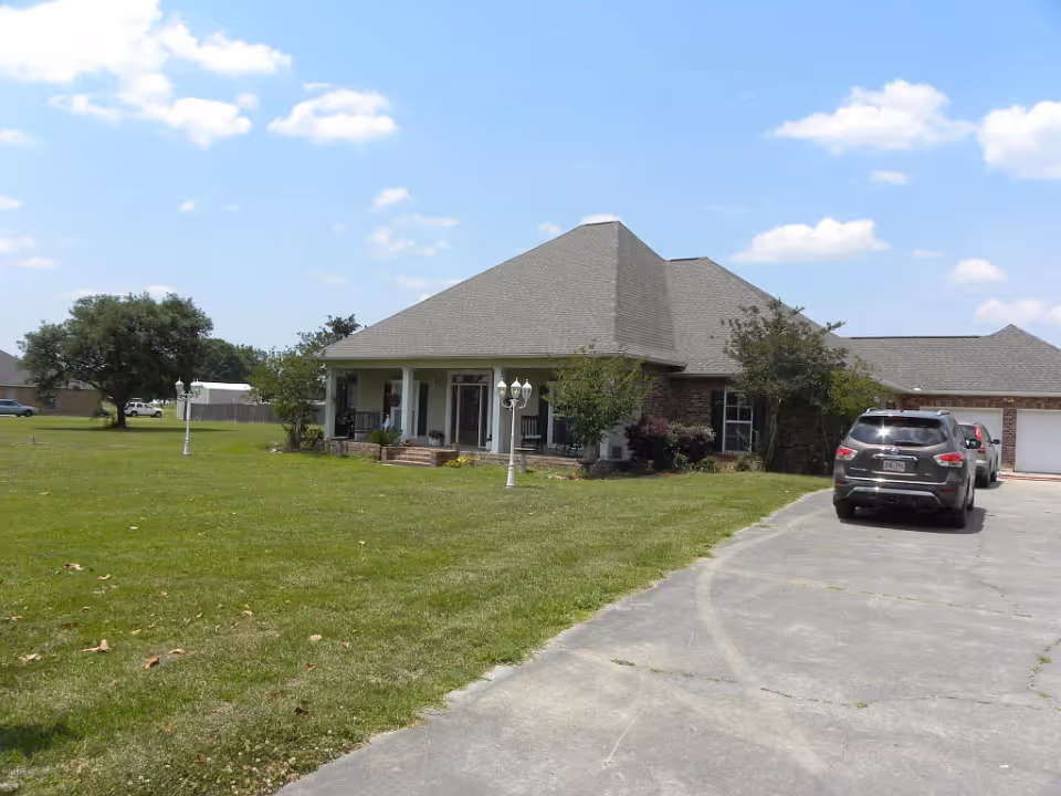 Front exterior of a single-story house with a large lawn, driveway, and parked cars under a blue sky.