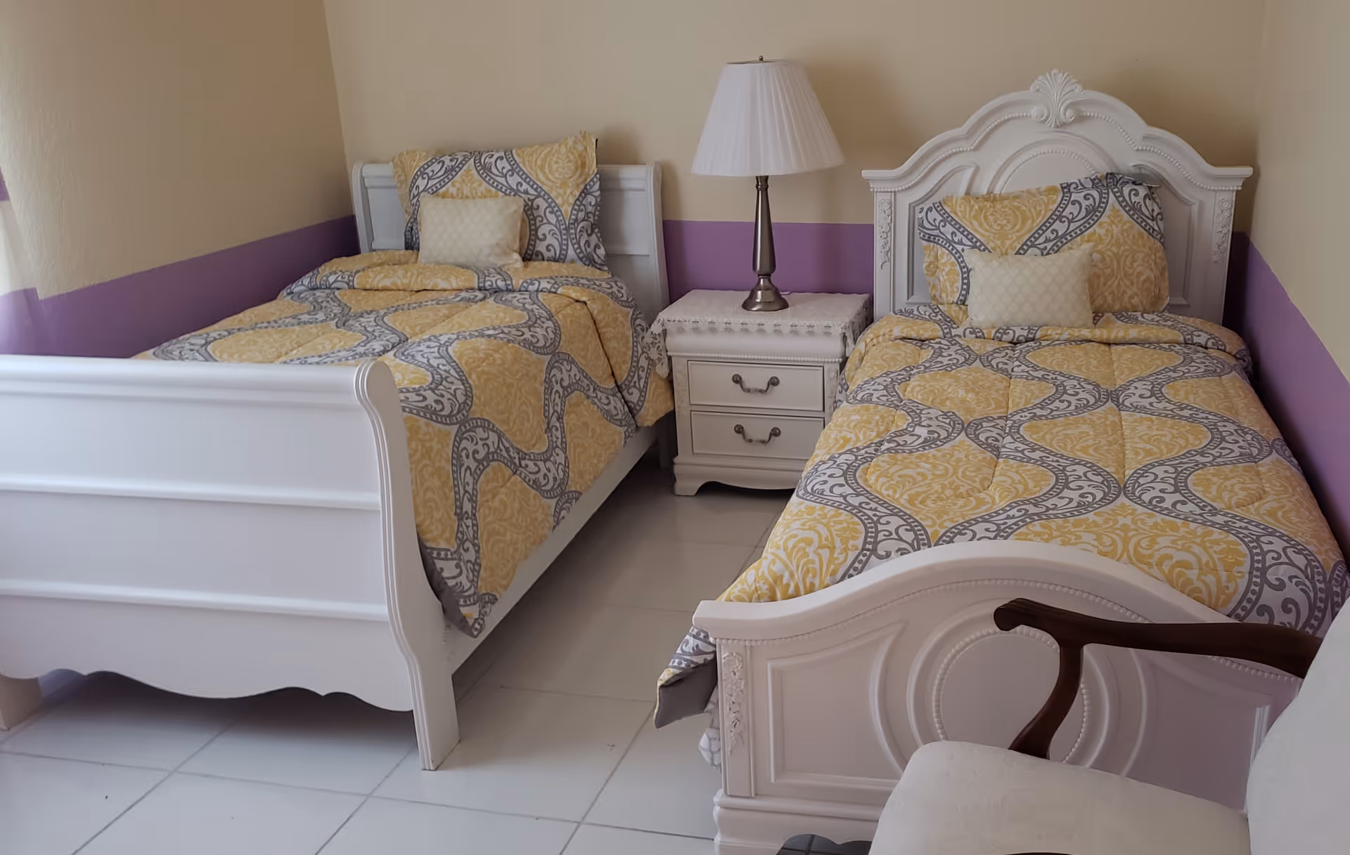 Two twin beds with yellow patterned bedding flanking a nightstand and lamp in a pastel-painted bedroom.
