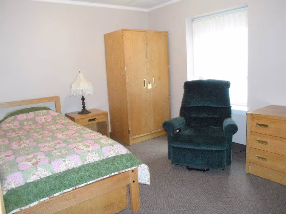 Simple bedroom with a single bed covered by a floral quilt, wooden furniture, a green recliner, and a wardrobe by a window.