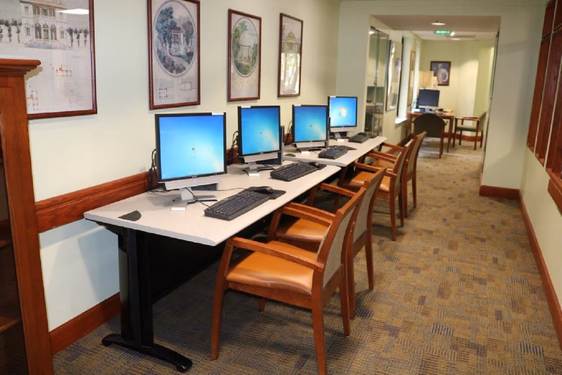 A computer lab area in a senior living facility with a row of four desktop computers on a long table, each accompanied by a wooden chair with padded seat. The walls are decorated with framed architectural drawings, and the carpet has a patterned design. The space is well-lit and has a hallway extending further back with additional computer stations.