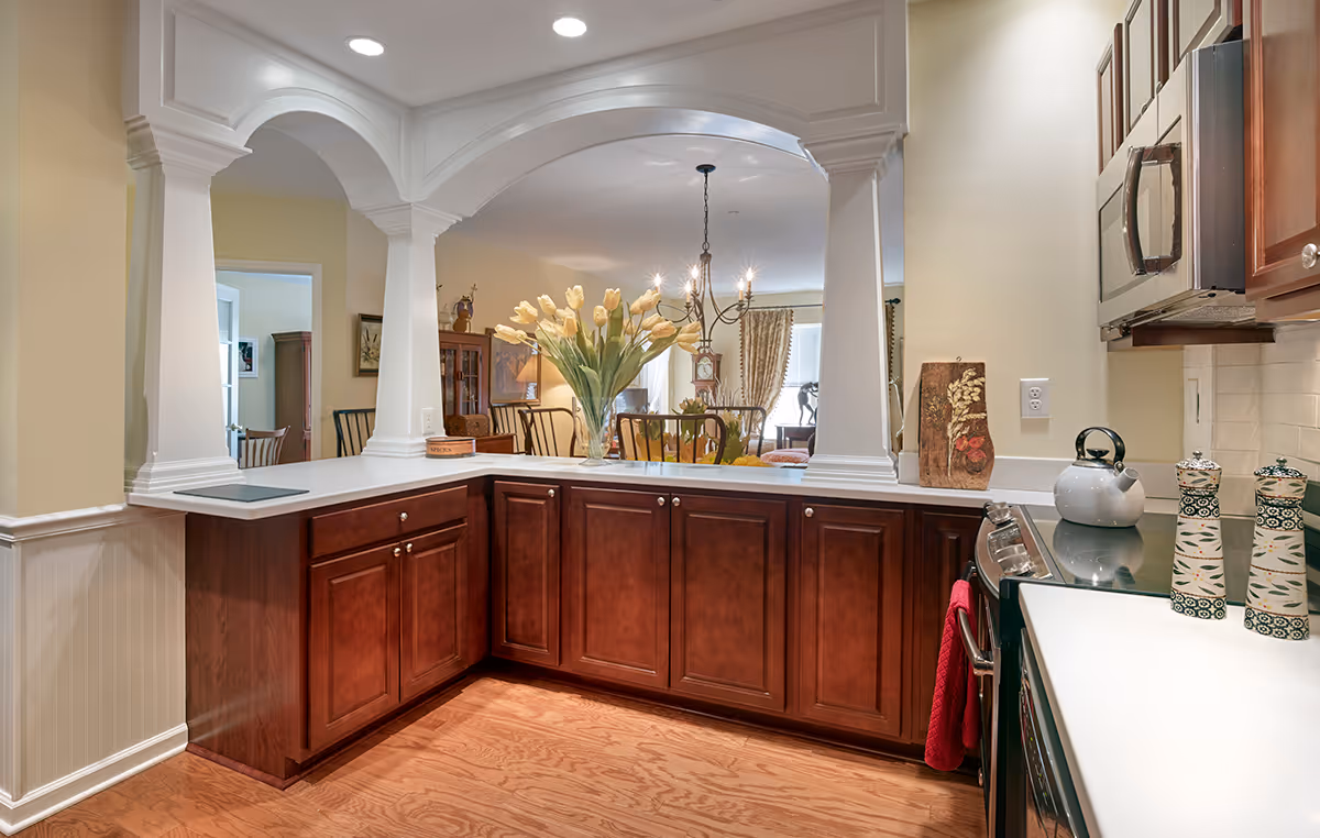 A bright kitchen with wooden cabinets and white countertops featuring a stove with a kettle, salt and pepper shakers, and a red towel. The kitchen opens to a dining and living area through a decorative white archway with columns. A vase with yellow tulips is placed on the counter, and the living area has chairs, a grandfather clock, and curtains on the window.