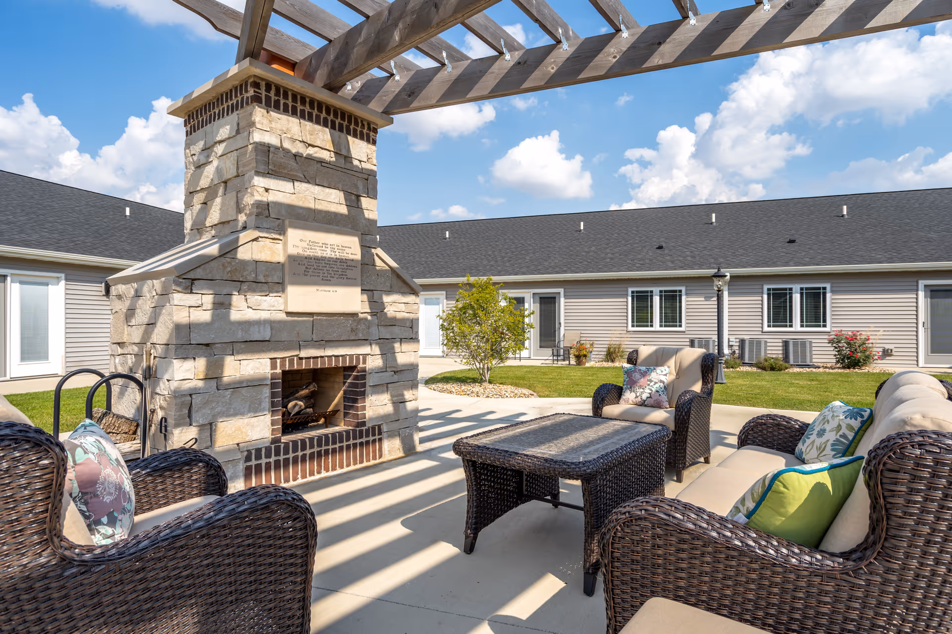 Sunlit outdoor courtyard featuring a stone fireplace under a pergola and wicker lounge seating in front of single-story residential units.