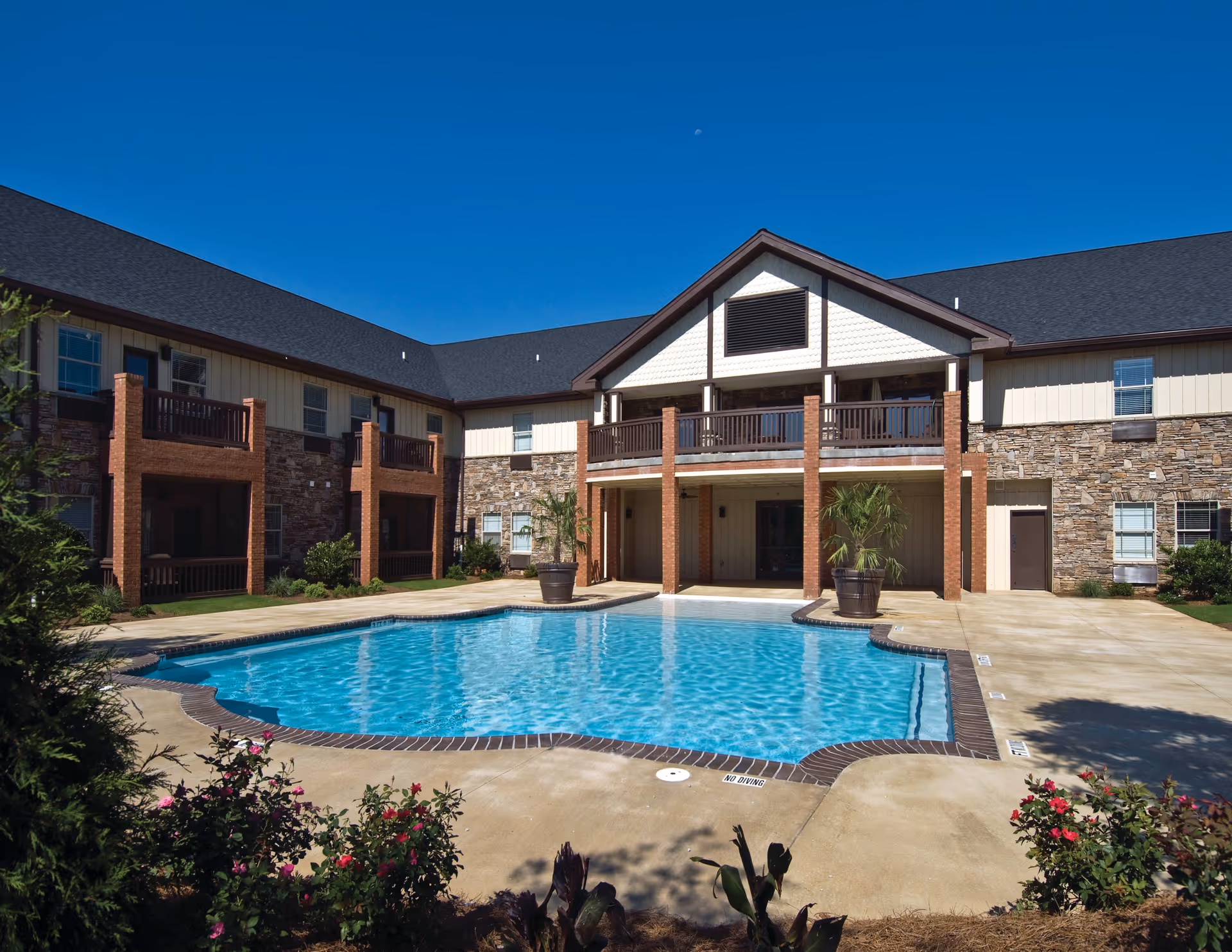 Outdoor swimming pool in the courtyard of a senior living facility with a two-story building surrounding it. The building has a mix of stone and siding exterior with balconies and a covered patio area. There are potted plants near the pool and flowering bushes in the foreground under a clear blue sky.