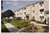 Three-story beige apartment-style building with a landscaped walkway and garden in front under a partly cloudy sky.