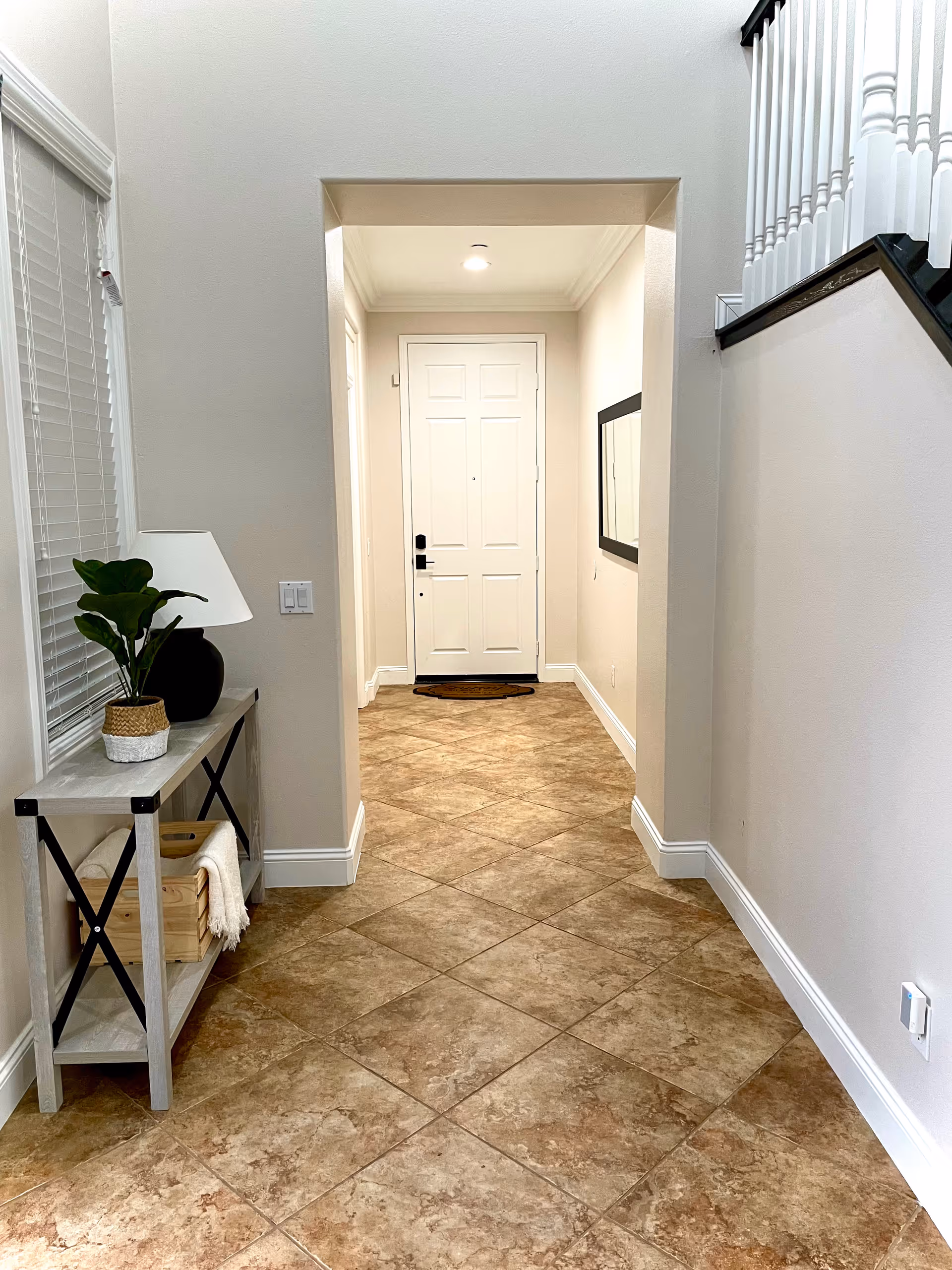 A hallway with beige tiled floor leading to a white front door. On the left side, there is a small console table with a potted plant, a lamp, and a wooden crate with a folded blanket. A large mirror is mounted on the right wall, and a staircase with white railings is partially visible on the right.