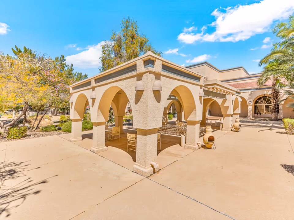 Outdoor patio area at Solstice Senior Living at Sun City West featuring a beige stucco pergola with arches, several chairs and tables underneath, surrounded by trees and plants under a bright blue sky with some clouds.