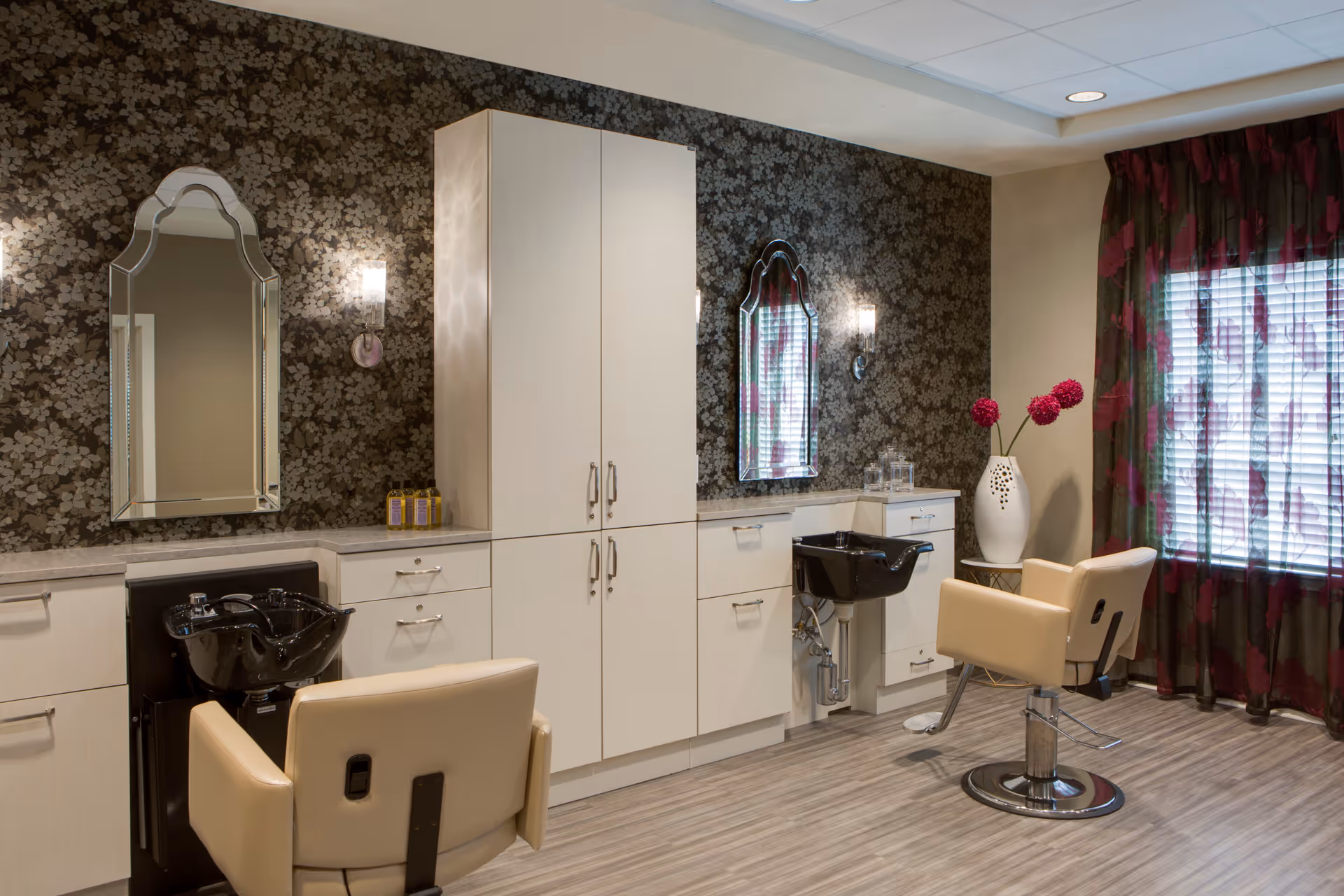 A hair salon area with two beige salon chairs facing black wash basins, white cabinetry, two decorative mirrors on a floral patterned wall, and a window with sheer curtains featuring a red floral design. A white vase with red flowers is placed near the window.