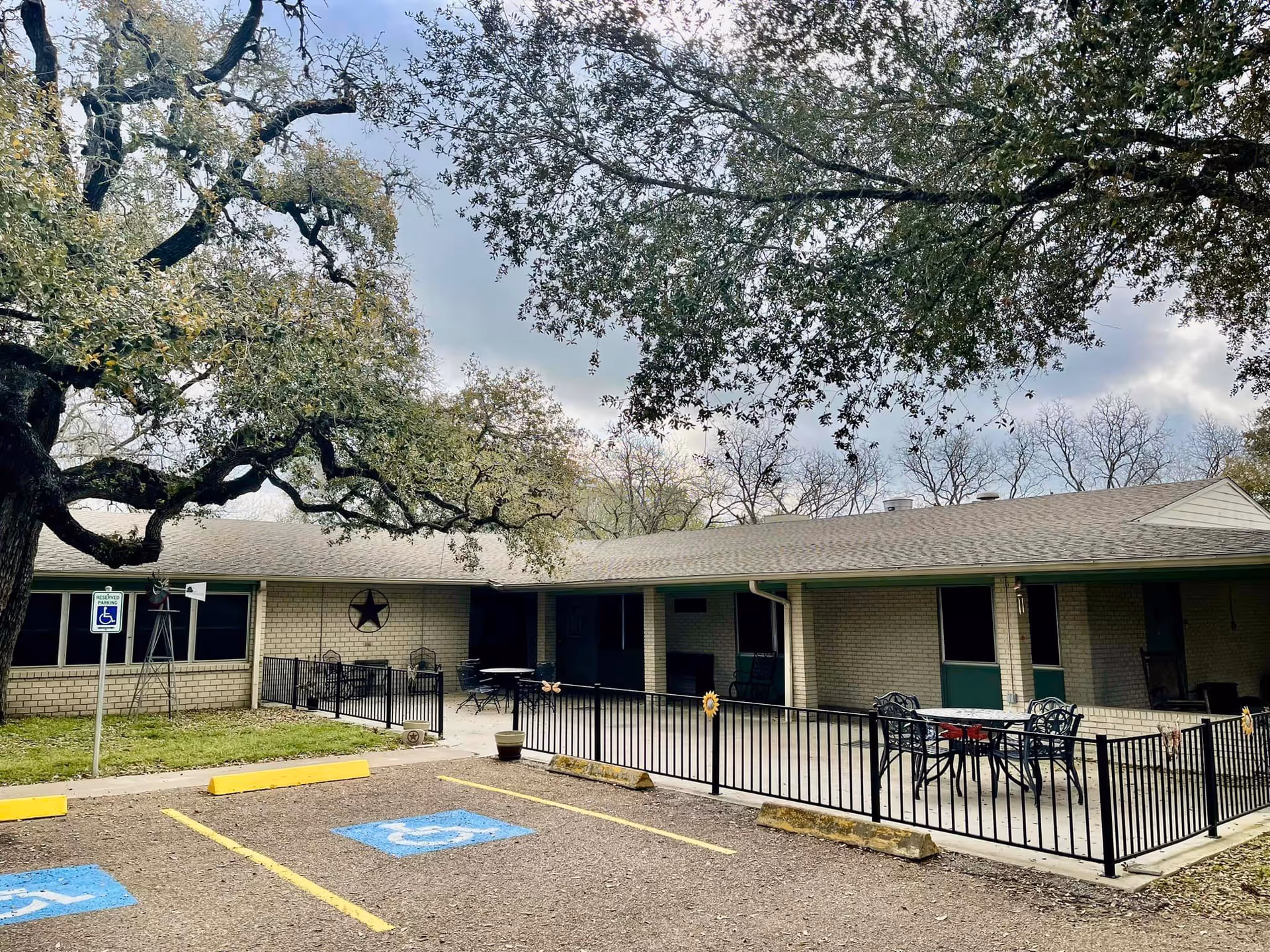 Exterior view of a single-story brick building with a covered patio area featuring outdoor tables and chairs. There are large trees with sprawling branches in the foreground and two handicap parking spaces marked in blue in front of the building. The sky is partly cloudy.