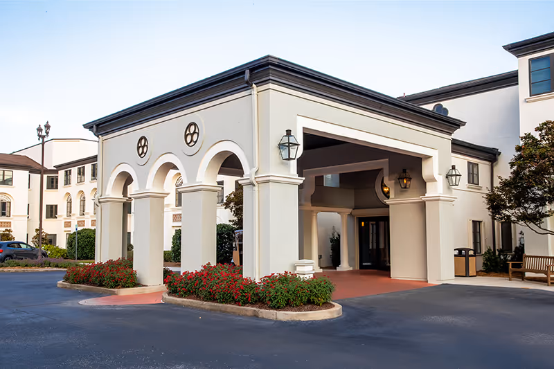 Entrance area of a senior living facility with a covered drop-off zone featuring arches and columns, surrounded by flower beds with red flowers, and a multi-story building in the background.