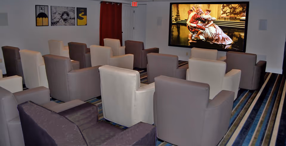 A small theater room with multiple rows of cushioned armchairs facing a large screen showing a scene from a movie. The room has striped carpeting, a white wall with three framed artworks, and a red curtain covering a door at the back.