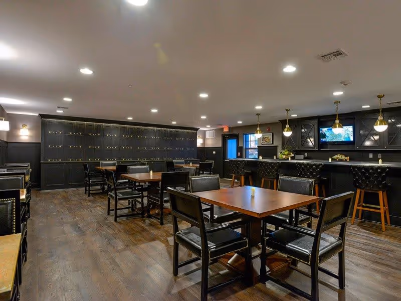 Interior view of a dining area with multiple wooden tables and black cushioned chairs arranged neatly on a wooden floor. A bar counter with high black cushioned stools is visible on the right side, with pendant lights hanging above it. The back wall features a large cabinet with multiple compartments and a mounted TV screen. The room is well-lit with recessed ceiling lights.