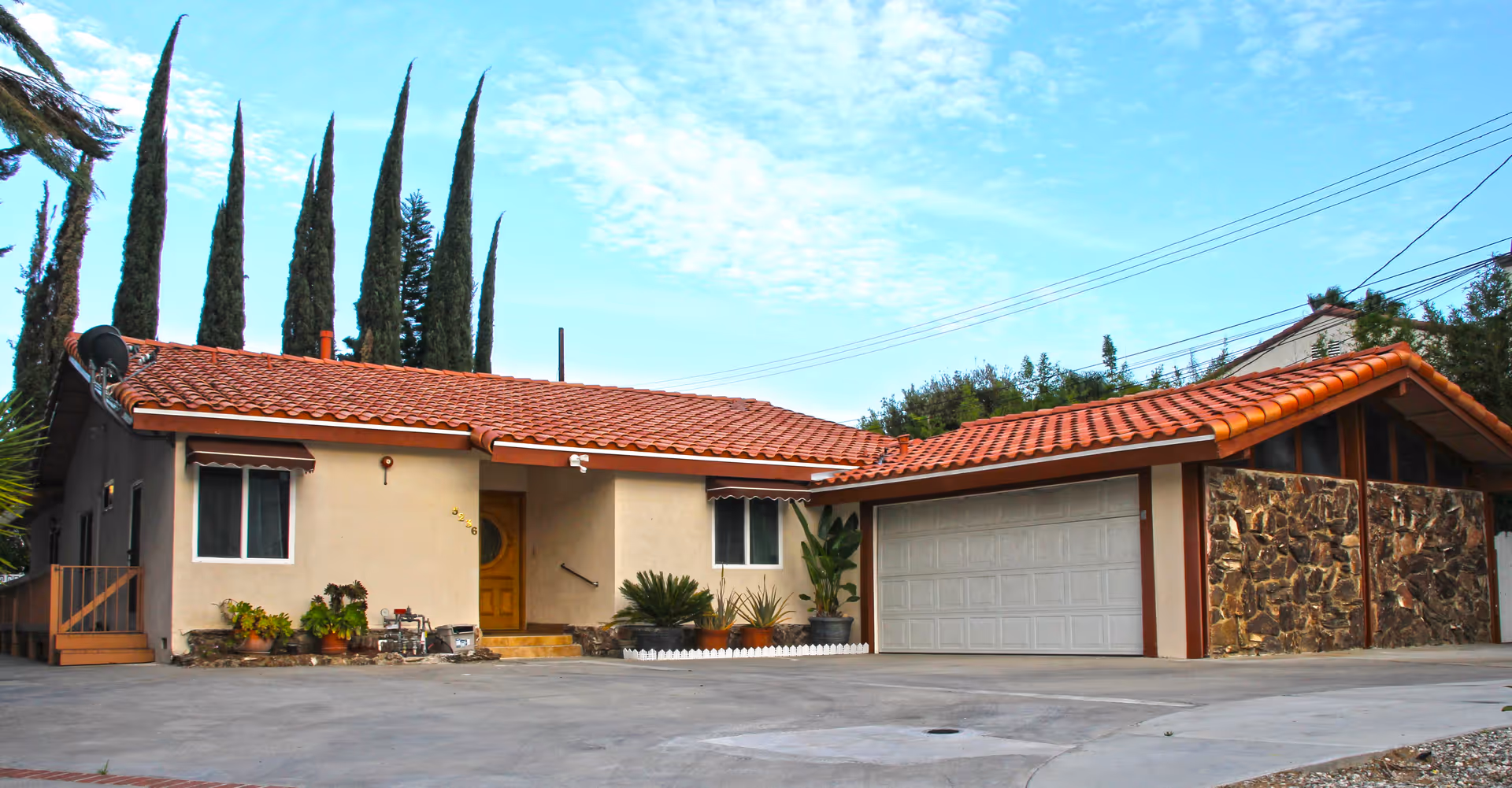 Exterior view of a single-story building with a red tile roof, beige walls, and a large garage door. Several tall, narrow trees are visible behind the building, and there are potted plants near the entrance. The sky is partly cloudy.