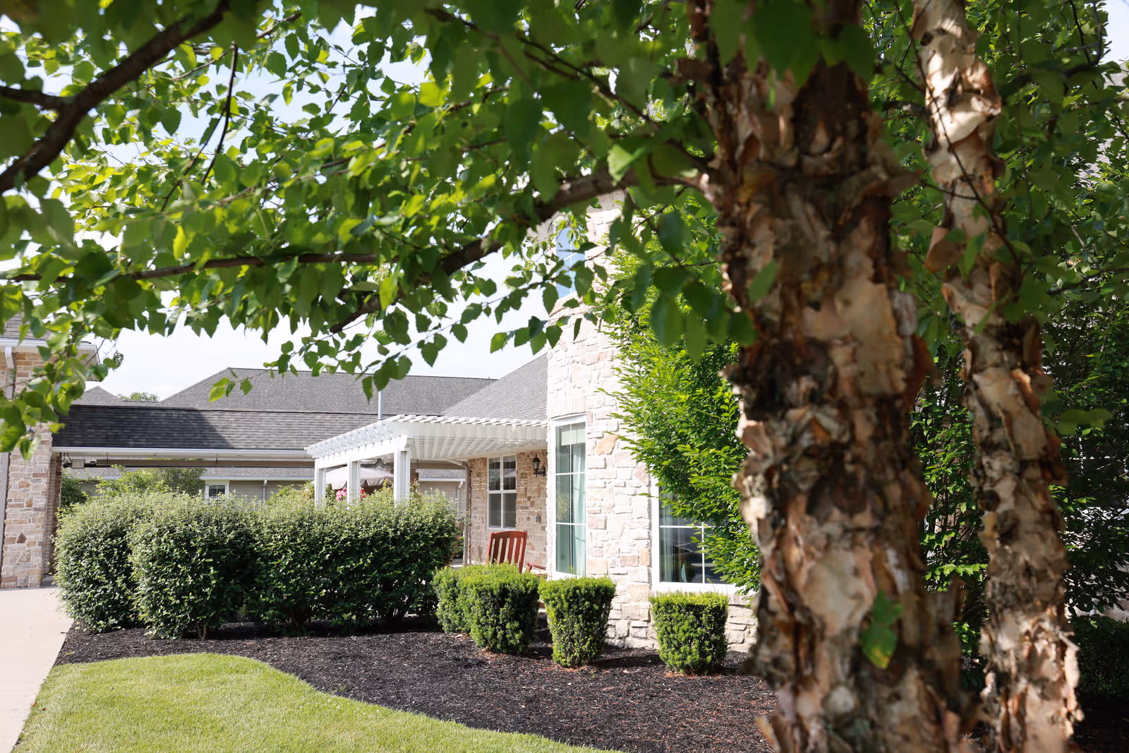 Front exterior of a senior living building with a porch, shrubs, and trees in the foreground.