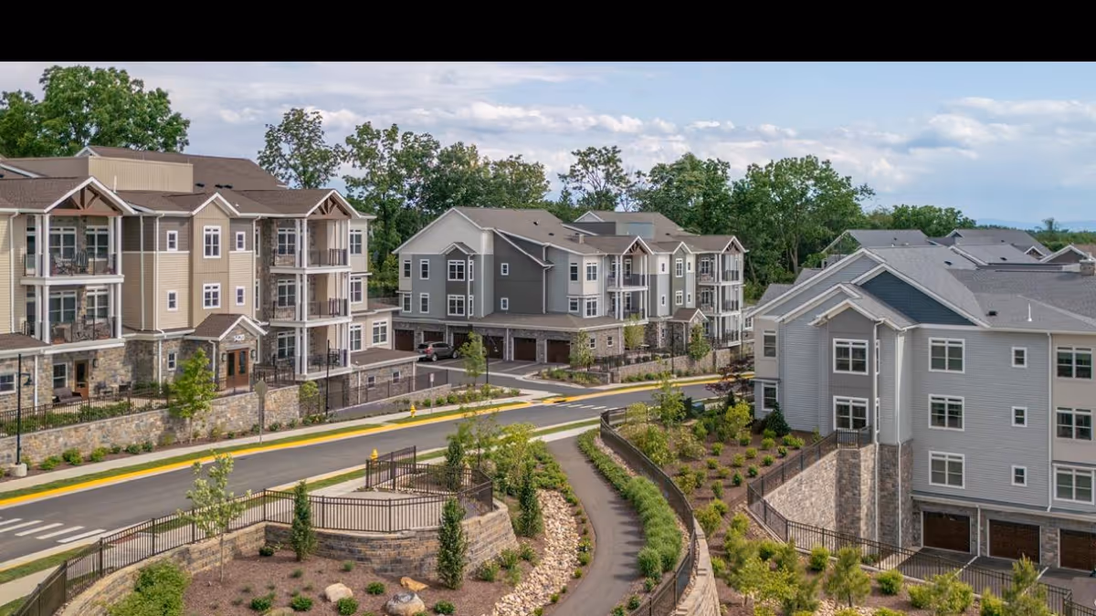 View of multiple modern residential buildings in a senior living community with landscaped gardens, paved roads, and trees under a partly cloudy sky.