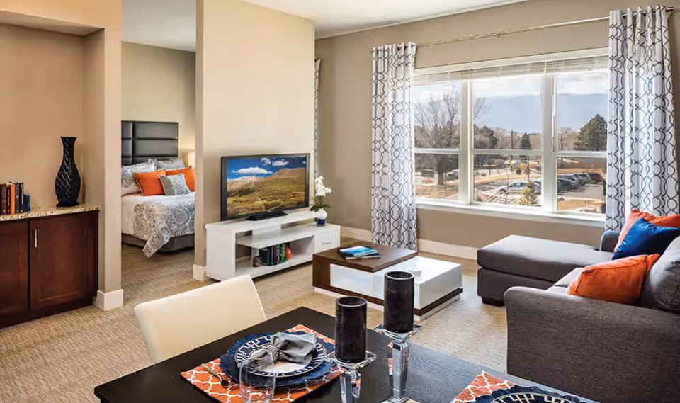 A bright and modern living room area with a large window showing an outdoor view. The room features a gray sectional sofa with orange and blue pillows, a white and wood coffee table, and a TV on a white stand. Adjacent to the living room is a dining table set with plates, glasses, and candles. A bedroom with a bed and pillows is visible through an open doorway. The walls are painted beige, and the floor is carpeted.