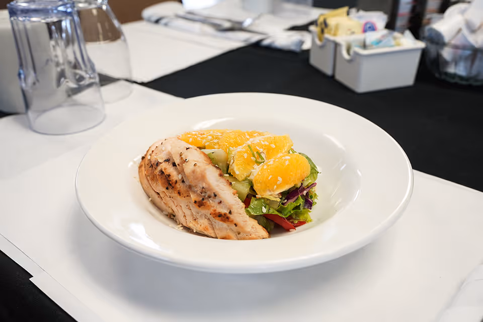 A white plate with a grilled chicken breast served alongside a fresh salad topped with orange slices and sesame seeds, placed on a table set with glasses, napkins, and condiment containers.