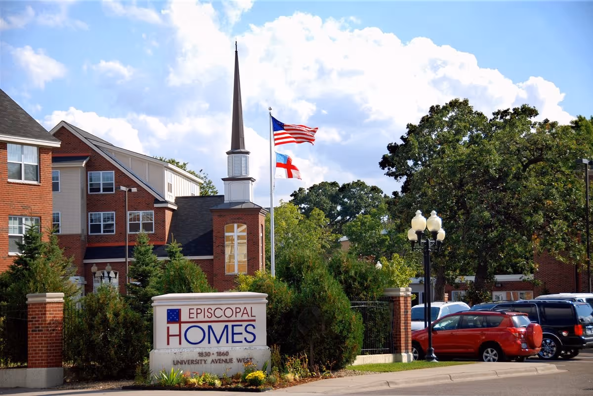 Brick senior living facility entrance with a sign reading 'Episcopal Homes', flags on a pole, landscaping, and parked cars.