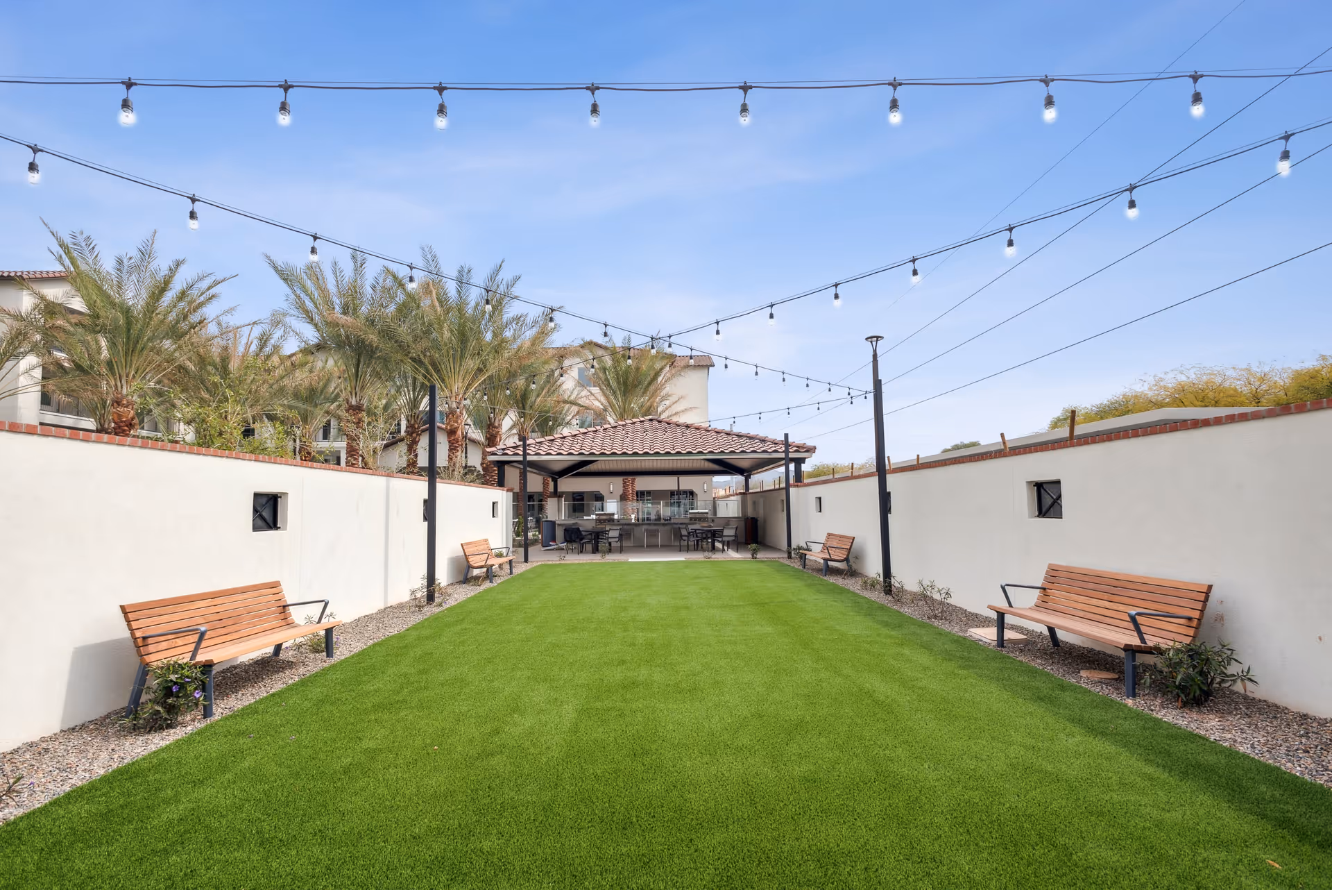Outdoor courtyard area with green artificial grass, four wooden benches along white walls on each side, string lights hanging overhead, palm trees in the background, and a covered patio with tables and chairs at the far end.
