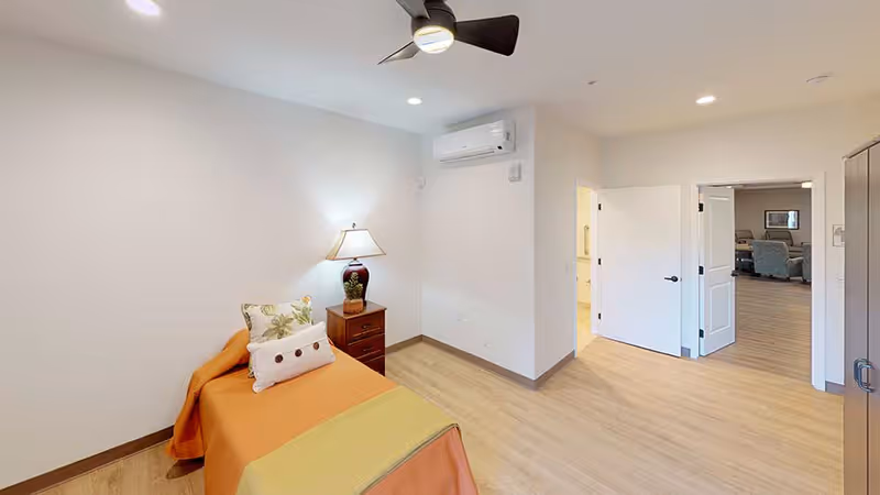 Small bedroom with a single bed draped in orange bedding, a nightstand with a lamp, ceiling fan and an open door to a living area.