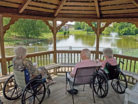 Three elderly individuals, two in wheelchairs and one seated in a chair, sitting under a wooden gazebo overlooking a pond with a fountain and greenery in the background.