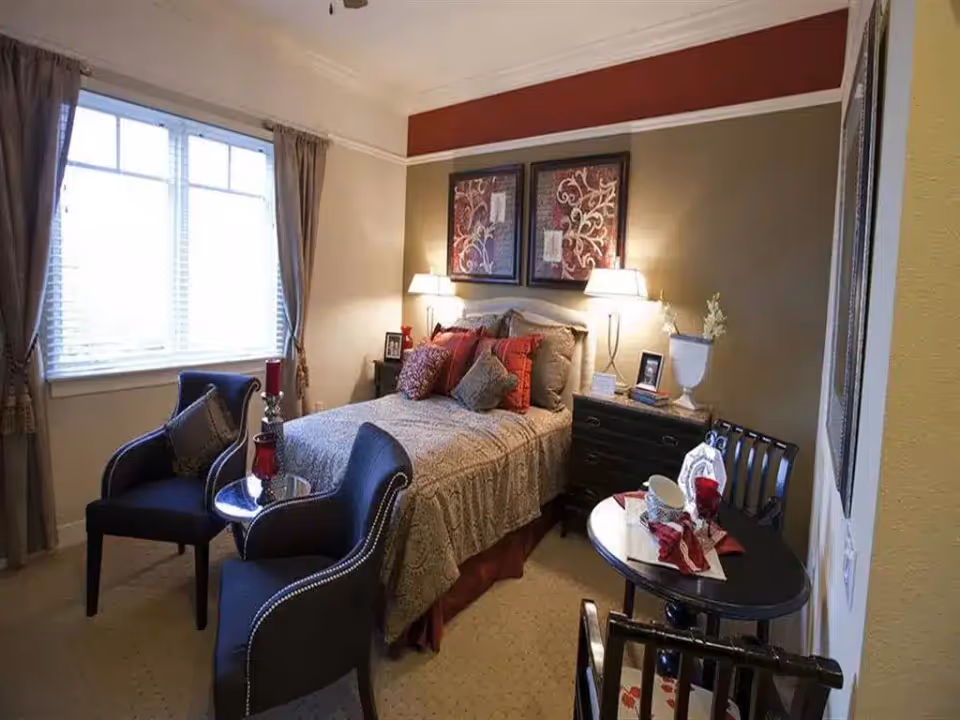 Cozy decorated bedroom with a bed, two armchairs, bedside tables with lamps, artwork and a small table near a window.