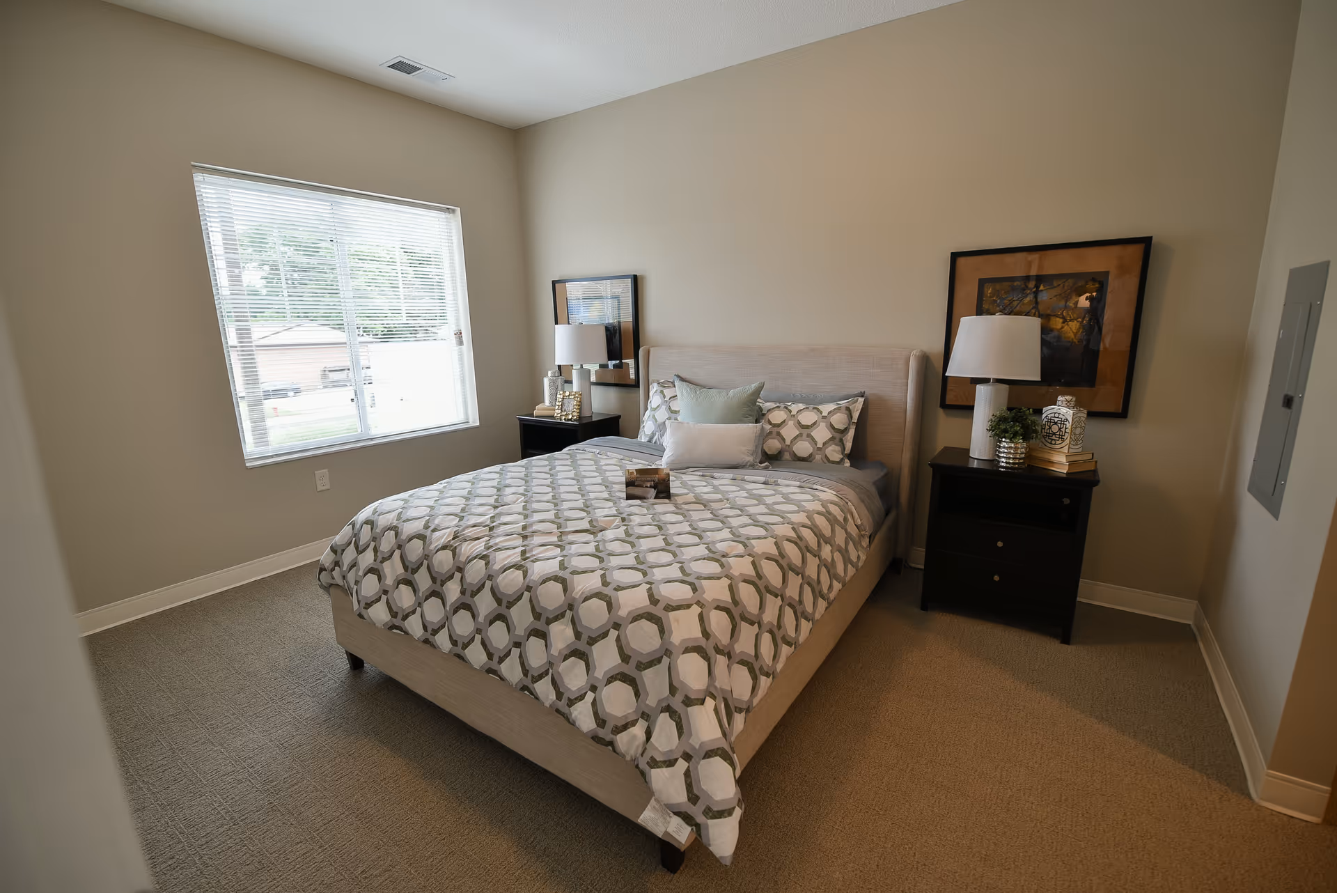 A bedroom with a large bed covered in a patterned comforter and multiple pillows. There are two black nightstands on either side of the bed, each with a lamp and decorative items. A window with blinds is on the left wall, and framed artwork hangs above each nightstand. The room has beige walls and carpeted flooring.