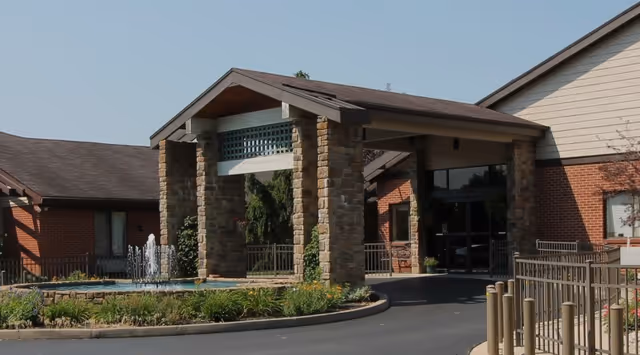 Entrance of a senior living facility with a covered driveway supported by stone pillars, a small water fountain surrounded by greenery, and a brick building under a clear blue sky.