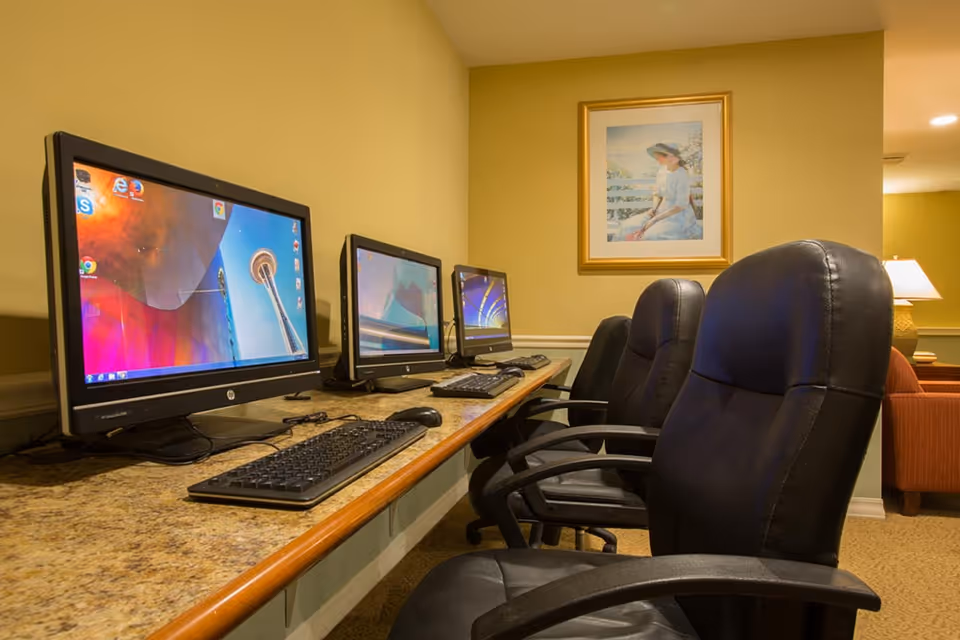 A computer workstation area with three desktop computers on a long granite countertop. Each computer has a keyboard and mouse, and there are three black leather office chairs in front of the computers. The walls are painted yellow, and a framed painting of a woman sitting outdoors hangs on the wall. In the background, there is a lamp and part of a red armchair visible.