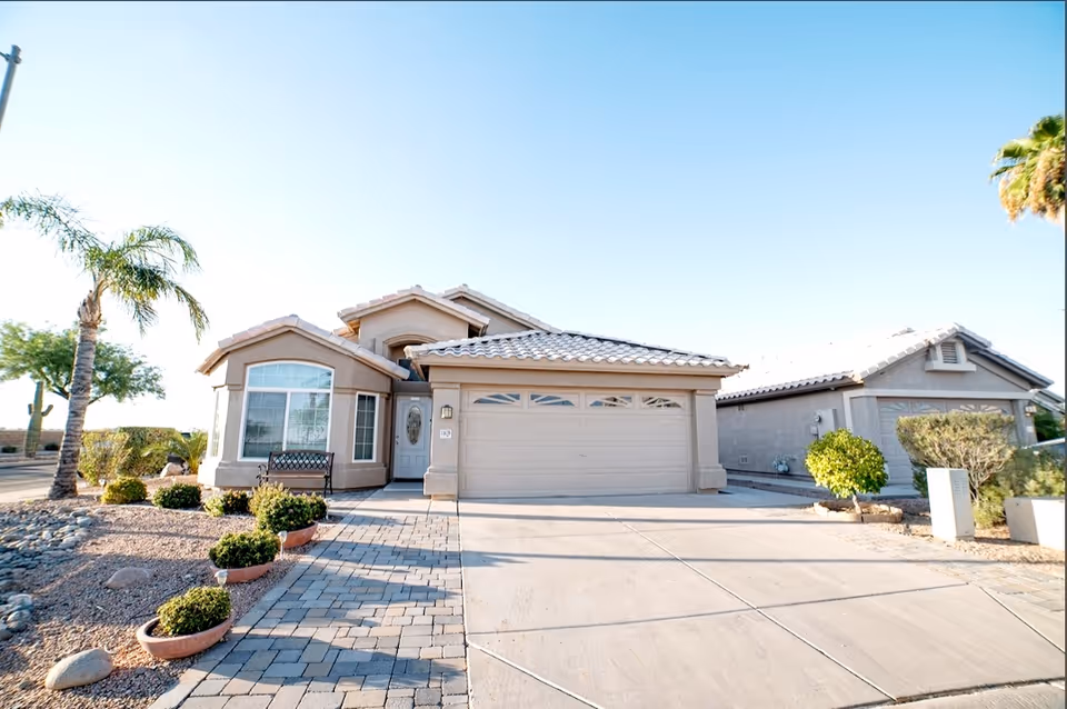 Front exterior of a single-story house with a two-car garage, driveway, and desert landscaping with palm trees.
