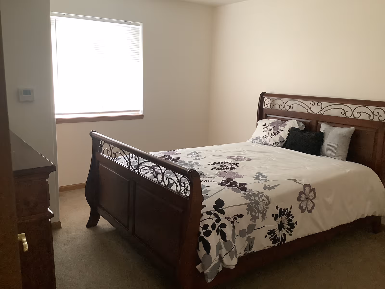 A bedroom with a wooden bed frame featuring decorative metalwork. The bed is made with a white comforter adorned with purple and gray floral patterns and several pillows. There is a window with blinds letting in natural light, and a wooden dresser is partially visible on the left side of the image.