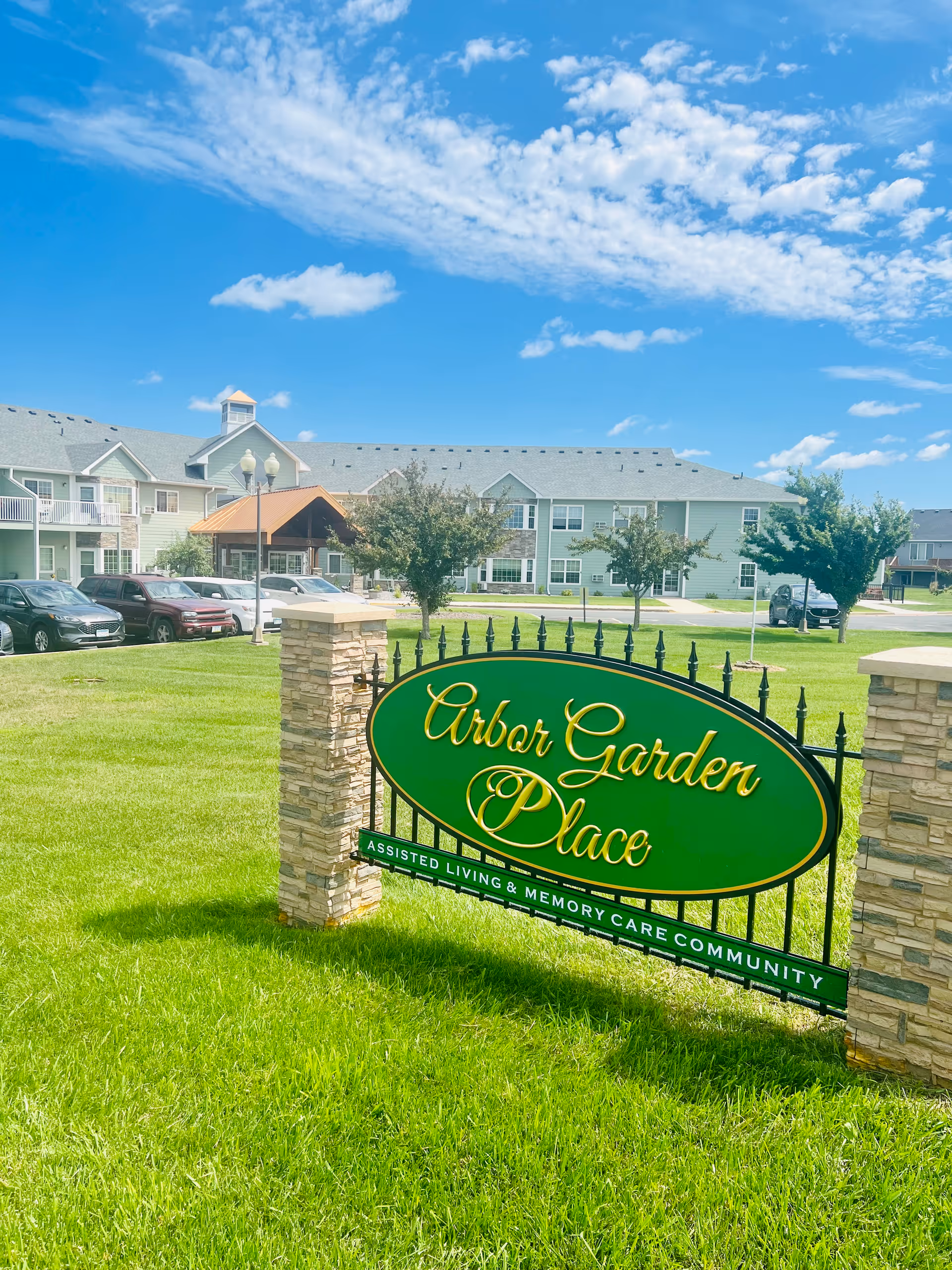 Outdoor view of Arbor Garden Place assisted living and memory care community sign on a green lawn with a multi-story residential building and parked cars in the background under a partly cloudy blue sky.