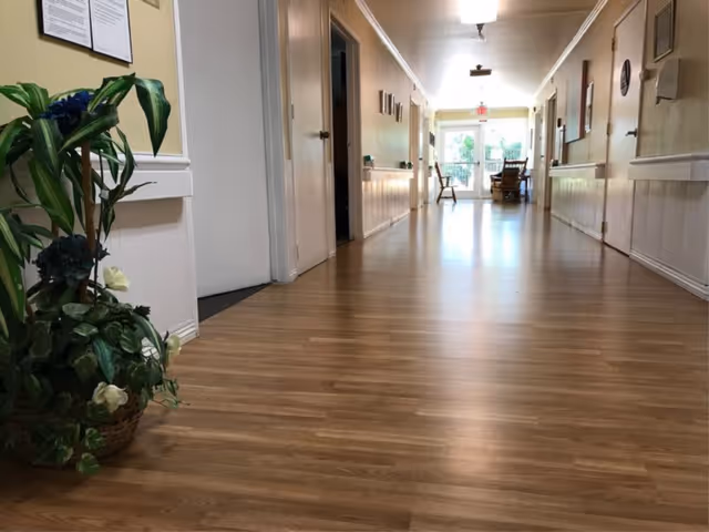 A long, well-lit hallway with wooden flooring in a senior living facility. There are doors on both sides of the hallway, a potted plant with green leaves and flowers on the left side near the foreground, and chairs visible at the far end near a glass door leading outside.