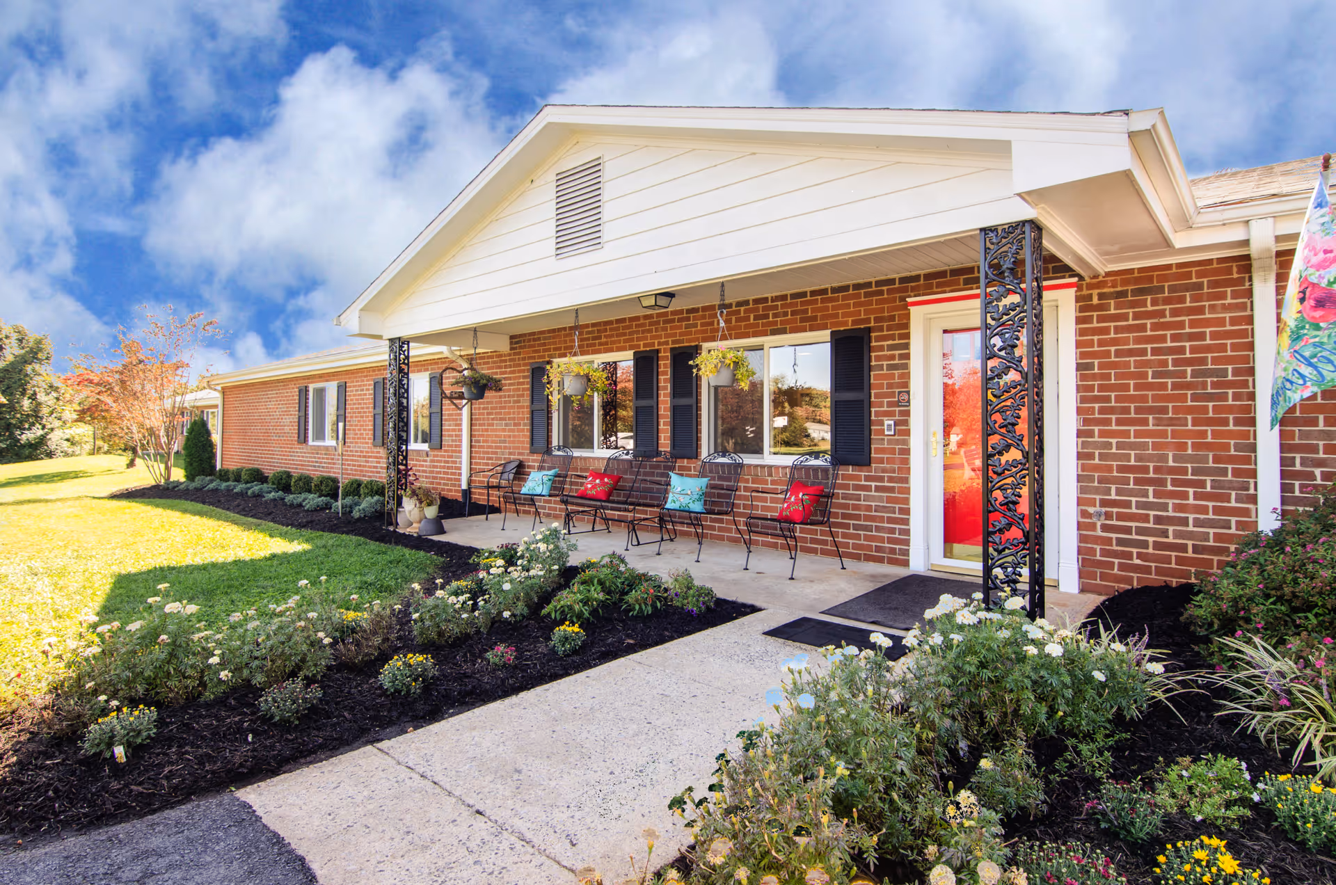 Front exterior view of a single-story brick building with a covered porch featuring black metal chairs with colorful cushions and hanging flower pots. The porch is supported by decorative black metal columns. There is a concrete walkway leading to the entrance door, surrounded by well-maintained flower beds and green grass under a partly cloudy blue sky.