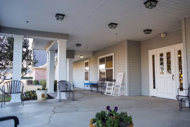 Covered outdoor porch area of a senior living facility with white columns, several metal chairs, a white rocking chair, and potted plants. The building has light gray siding and white double doors with glass panels.
