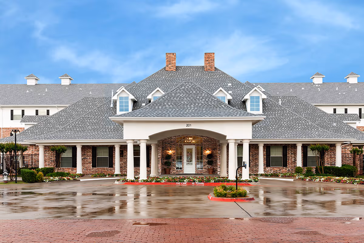 Front entrance of a brick senior living building with white columns, pitched roof dormers, and a wet driveway.