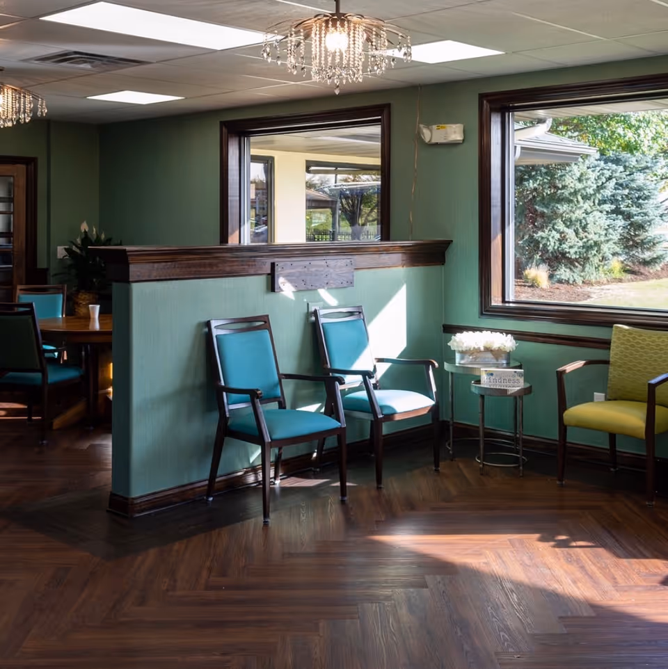 Sunlit communal sitting area with teal and green chairs, wood flooring, large windows, and a small side table with flowers.