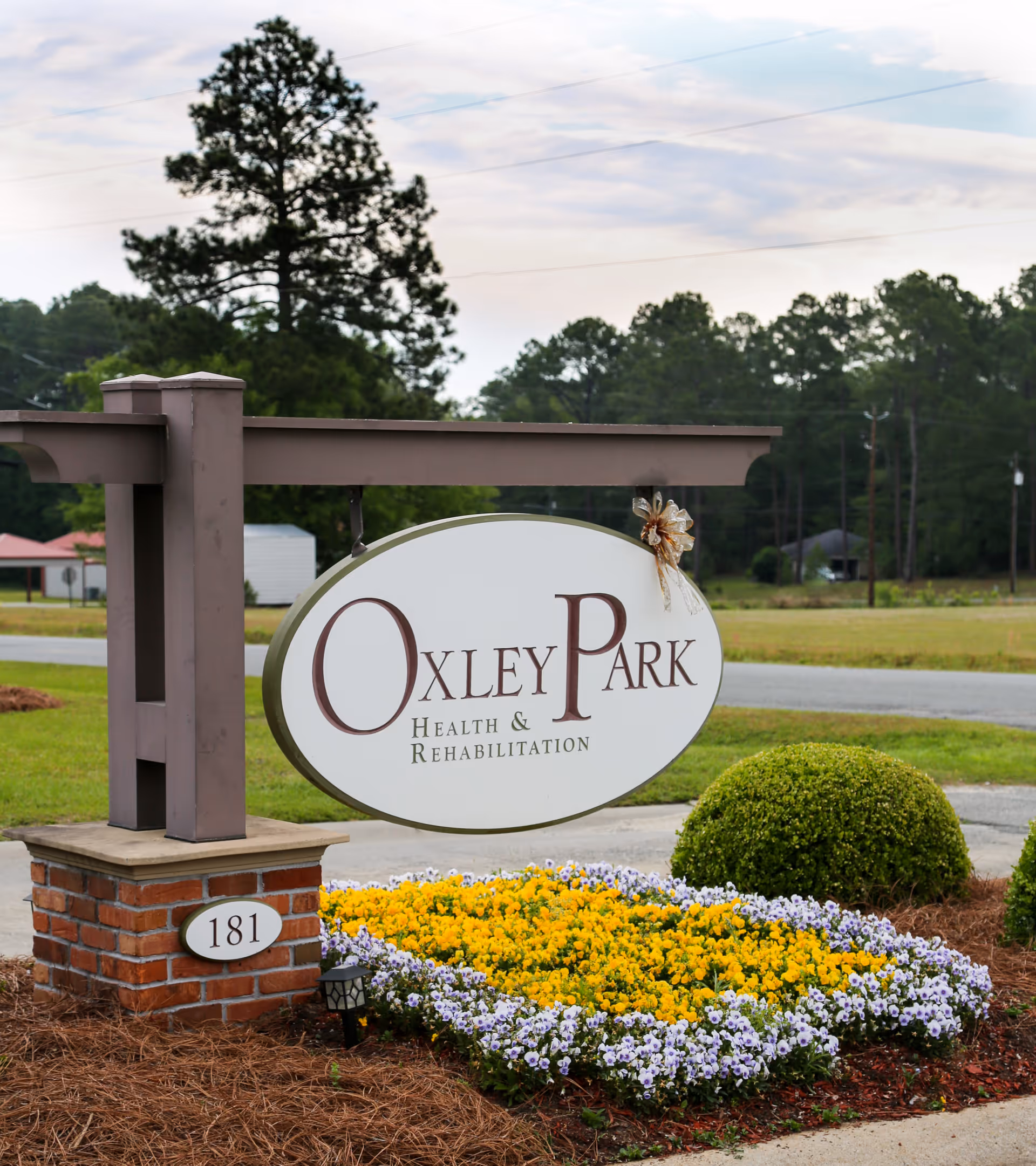Outdoor sign for Oxley Park Health & Rehabilitation mounted on a wooden post with a brick base, surrounded by a flower bed with yellow and purple flowers, green bushes, and a road and trees in the background.