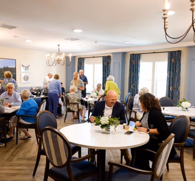 A bright dining room with several elderly people and staff members interacting and enjoying their time. The room has round tables with chairs, floral centerpieces, large windows with blue patterned curtains, and chandeliers hanging from the ceiling.