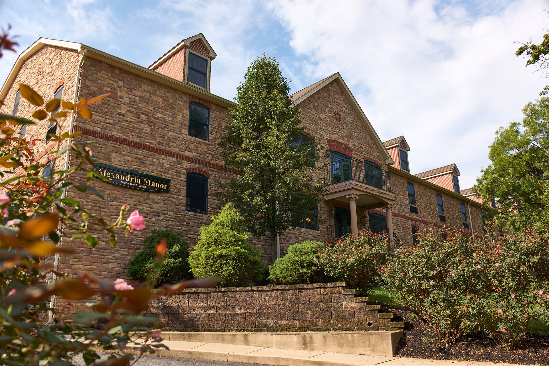 Exterior view of Alexandria Manor, a senior living center, showing a large stone building with multiple windows, a small balcony, and surrounded by greenery and flowering bushes under a partly cloudy sky.
