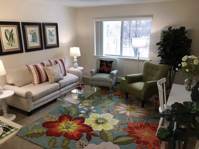 A cozy living room with a white sofa adorned with striped and patterned pillows, two armchairs—one green and one striped—, a glass coffee table on a colorful floral rug, two white side tables with lamps, framed botanical prints on the wall, a large window with a view of trees, and several green plants including a tall one in the corner.