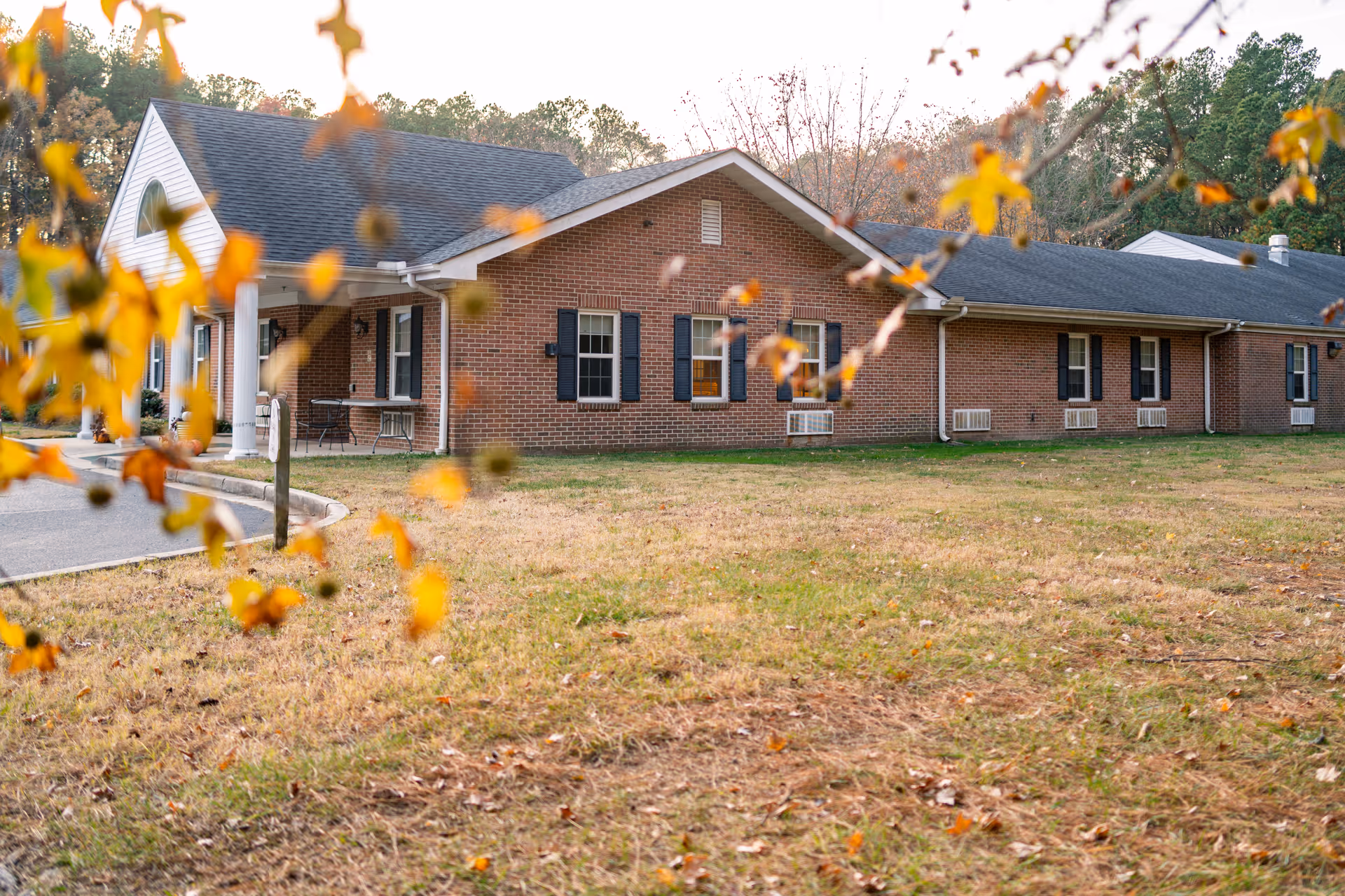Exterior view of a single-story brick building with black shutters and a dark shingled roof, surrounded by a grassy area with some autumn leaves in the foreground and trees in the background.