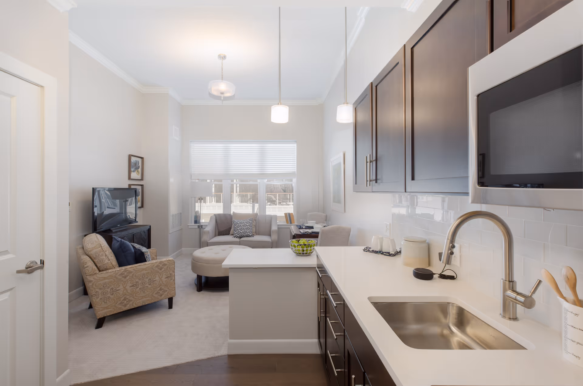 A bright and modern interior space featuring a small kitchen area with dark wood cabinets, a stainless steel sink, and a microwave. The kitchen opens into a cozy living room with a beige armchair, a small sofa, an ottoman, a TV on a stand, and a window with blinds letting in natural light.