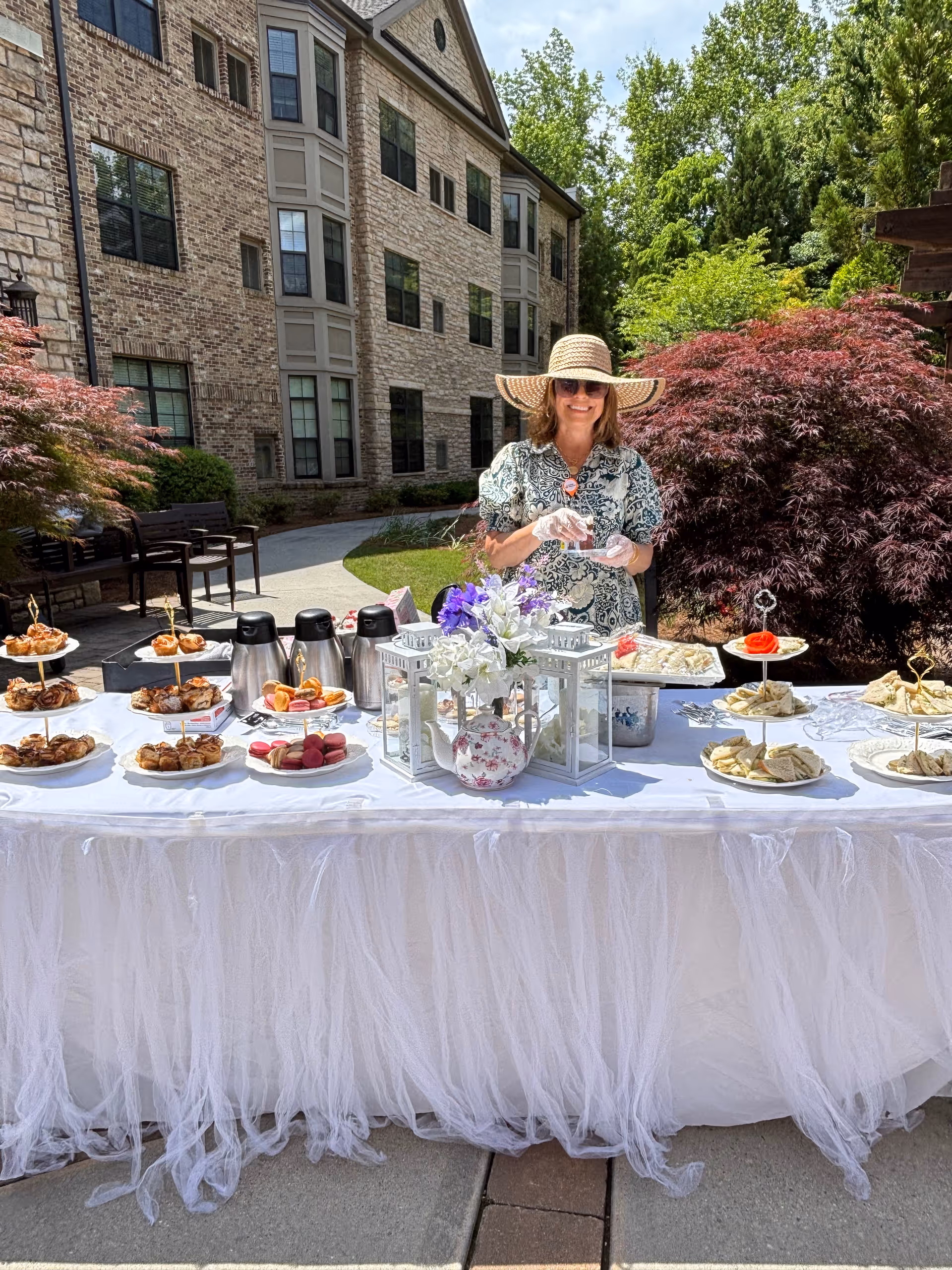 A woman wearing a wide-brimmed hat and sunglasses stands behind a long table covered with a white tablecloth and decorated with flowers and lanterns. The table is set outdoors with various pastries, sandwiches, and coffee pots. Behind her is a multi-story brick building and lush greenery.
